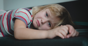 Child with blonde hair in a red striped shirt laying sideways on a sofa looking sad.